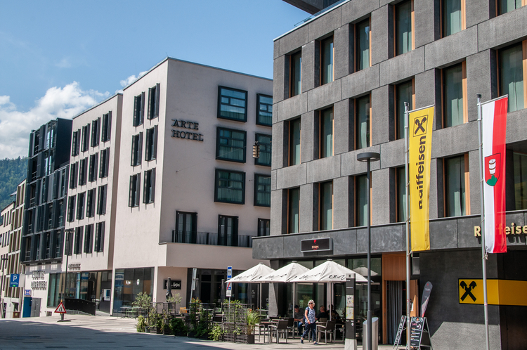 Eine städtische Szene zeigt das Arte Hotel an einer Straße mit modernen Gebäuden und einem Café im Freien. Die Fassade des Hotels ist hell mit großen Fenstern. Vor dem Café sind Tische und Stühle aufgestellt, und es wehen Fahnen in der Nähe.